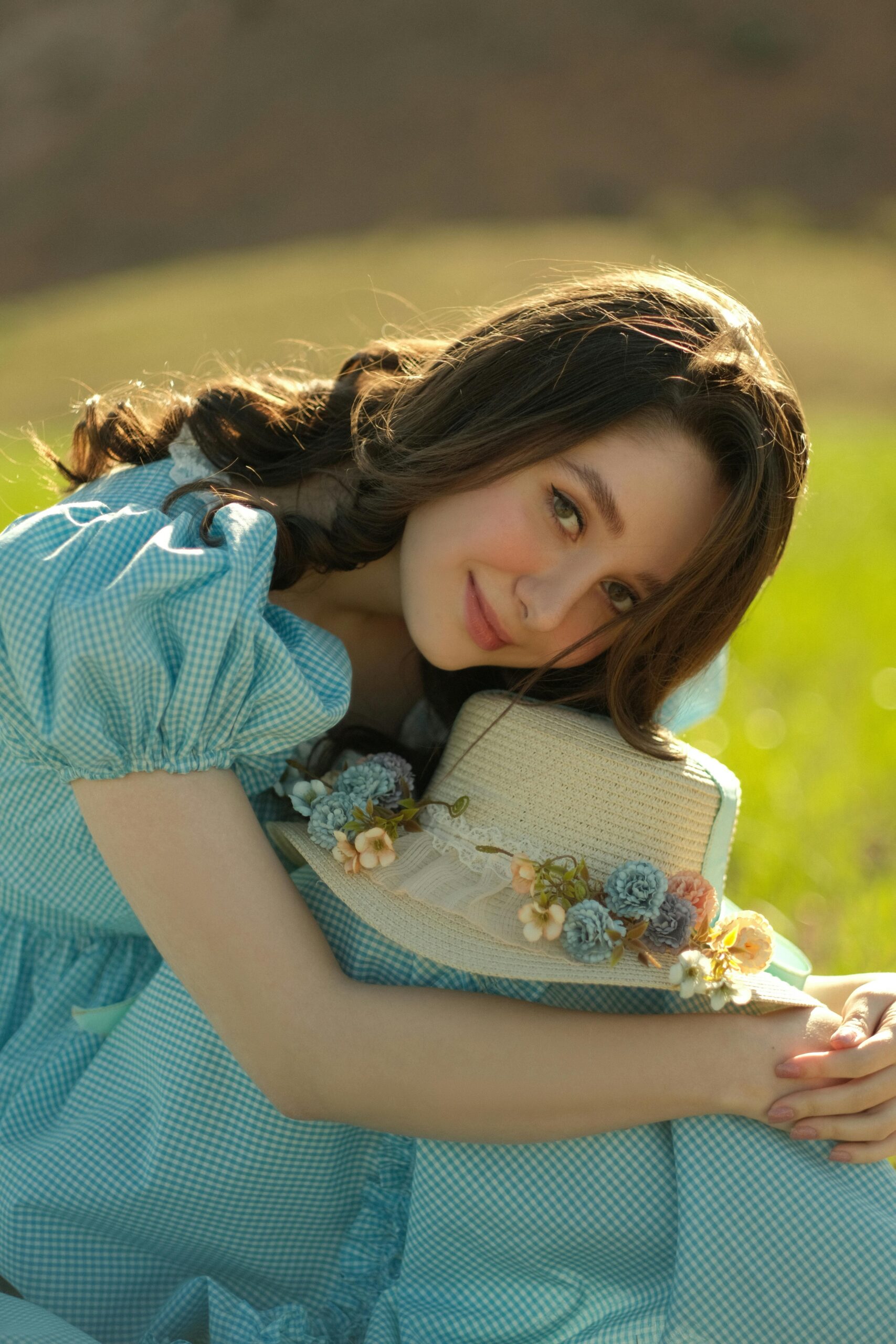 A young woman with brunette hair in a vintage blue dress poses outdoors with a floral hat.