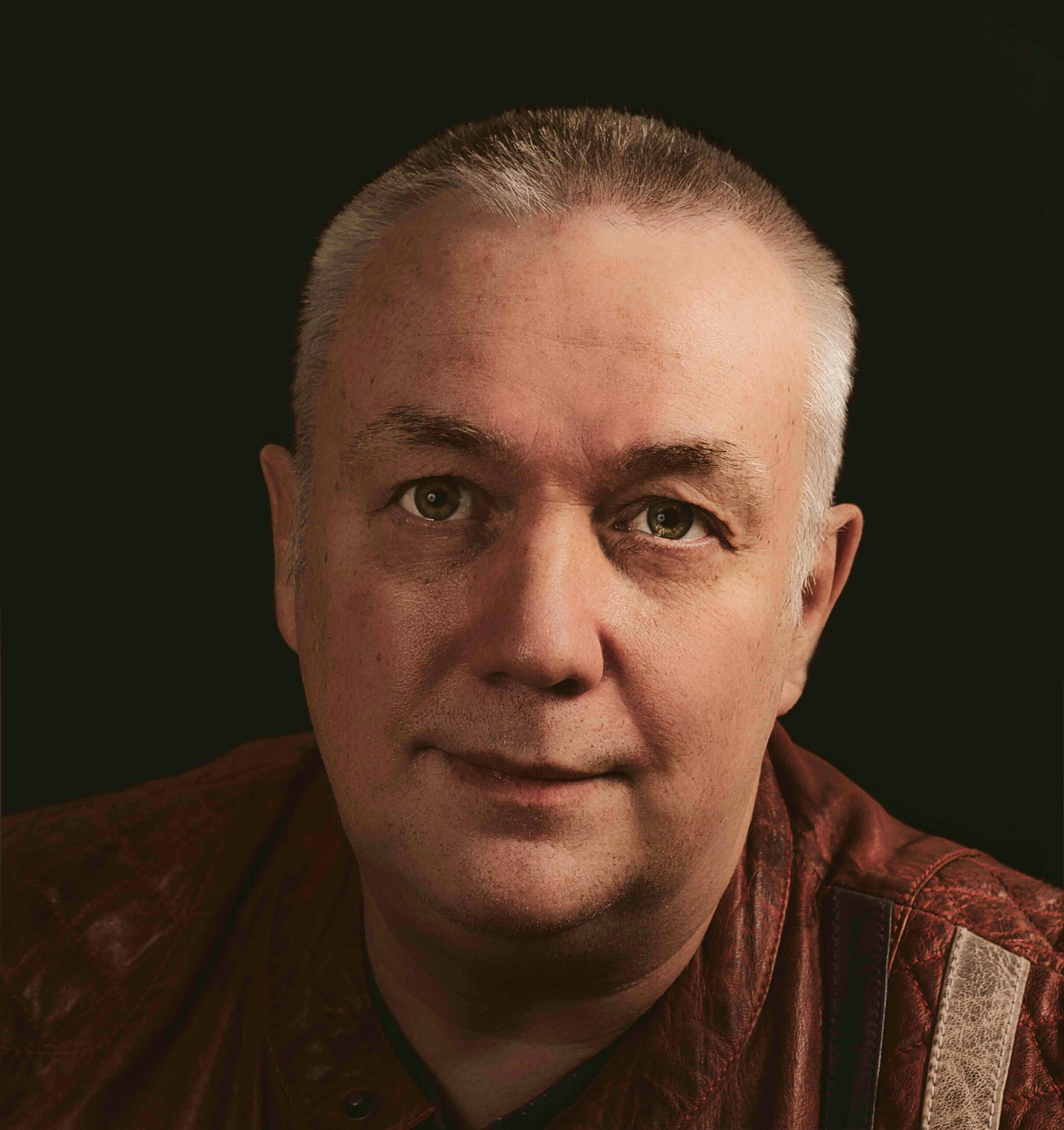 Portrait of content mature male with short haircut wearing brown shirt in studio against black background